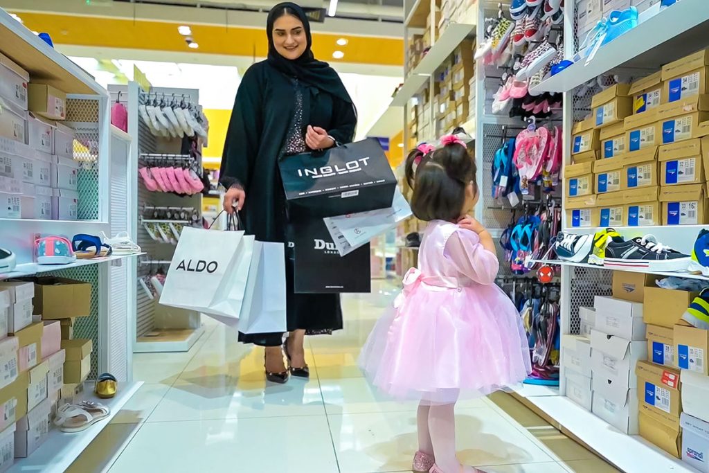 A little girl in a pink dress is standing in front of a shoe stand, next to her mother that carrying a lot of bags